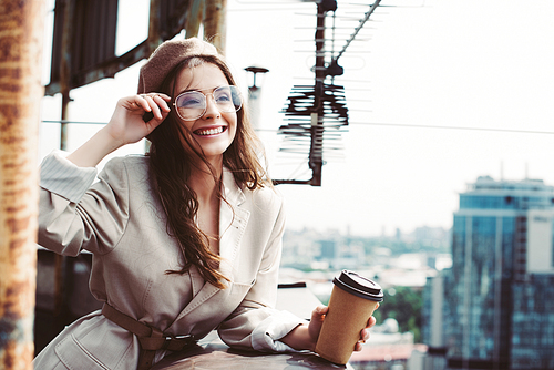 beautiful smiling girl in beige suit and beret posing on roof with coffee to go