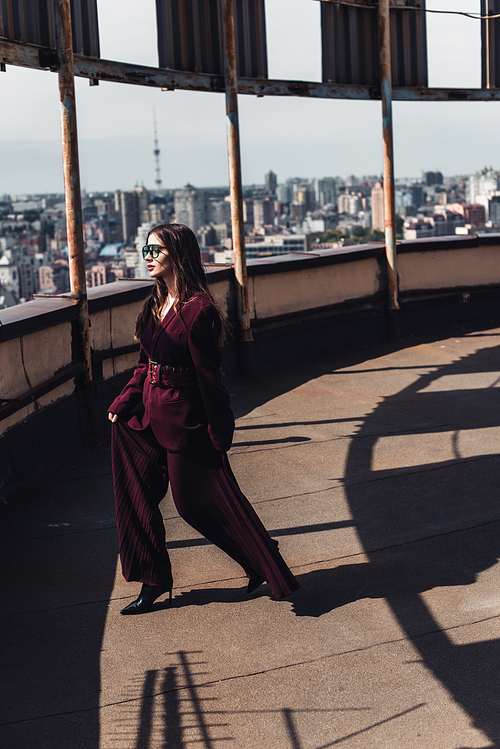 attractive elegant woman posing in trendy burgundy suit and sunglasses on urban roof