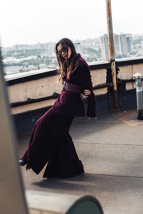 attractive elegant woman posing in trendy burgundy suit and sunglasses on urban roof