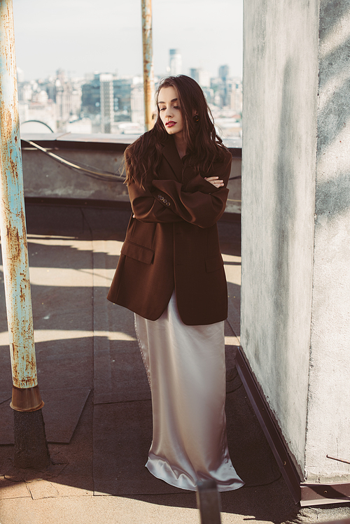 attractive fashionable woman posing in silk dress and brown jacket on roof