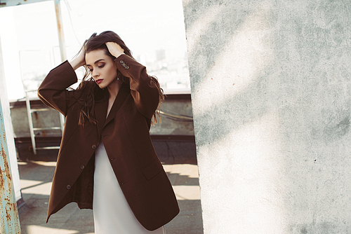 attractive stylish woman posing in silk dress and brown jacket on roof