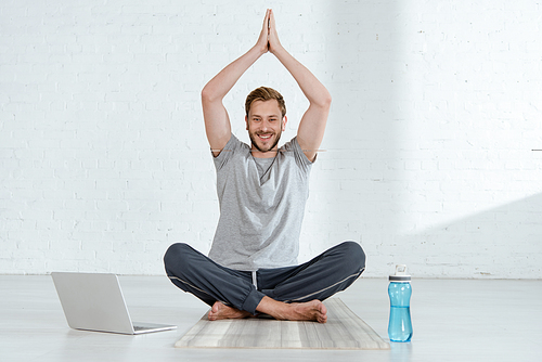 smiling man  while practicing half lotus pose with raised prayer hands near laptop and sports bottle