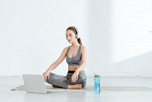 young woman in headphones sitting in easy pose near laptop and sports bottle