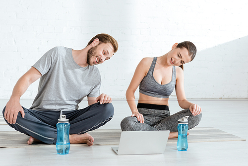 smiling man and woman looking at laptop while sitting in easy poses near sports bottles