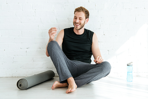 cheerful man sitting on floor near yoga mat and sports bottle and holding earphones