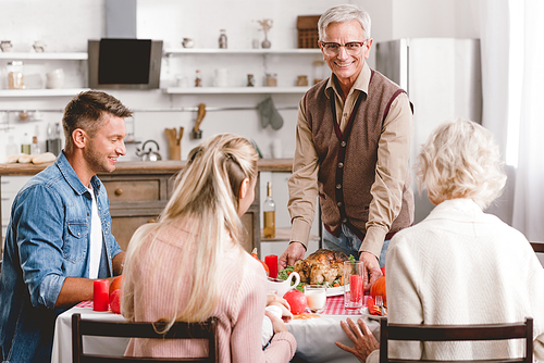 family members sitting at table and grandfather holding plate with turkey in Thanksgiving day