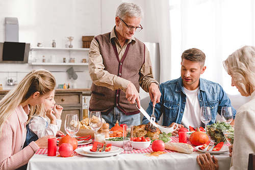 family members sitting at table and grandfather cutting tasty turkey in Thanksgiving day