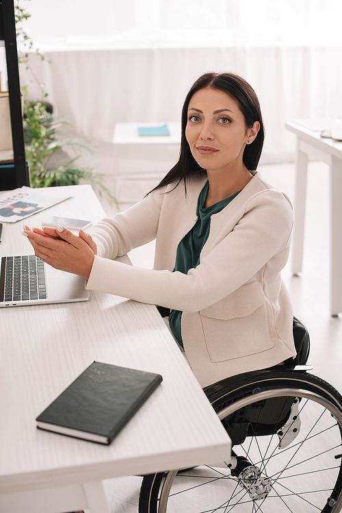 beautiful disabled businesswoman smiling at camera while sitting at workplace in wheelchair