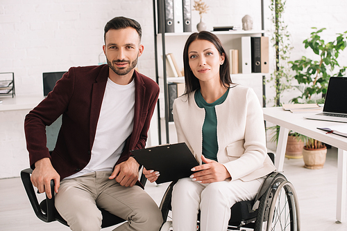 positive disabled businesswoman holding clipboard while sitting near business partner in office