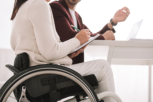 cropped view of disabled businesswoman writing in notebook while sitting near business partner
