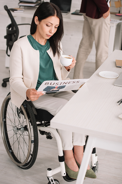 attractive disabled businesswoman reading newspaper and holding coffee cup while sitting in wheelchair at workplace