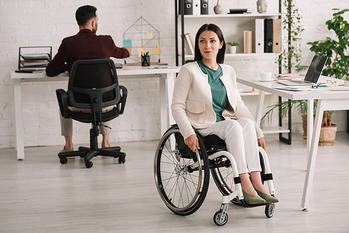 attractive disabled businesswoman sitting in wheelchair in office near colleague