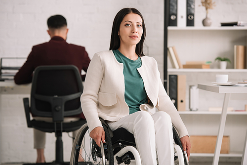 smiling disabled businesswoman  while sitting in wheelchair near colleague working on background