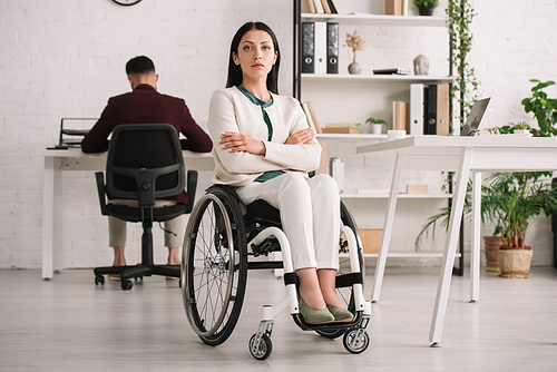 confident disabled businesswoman sitting in wheelchair with crossed arms near collegue working on background