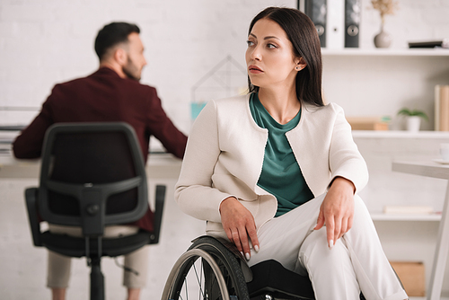 serious disabled businesswoman looking away while sitting in wheelchair in office near colleague