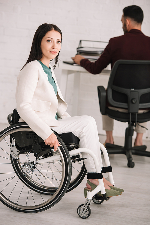 attractive disabled businesswoman smiling at camera while sitting in wheelchair near colleague working on background