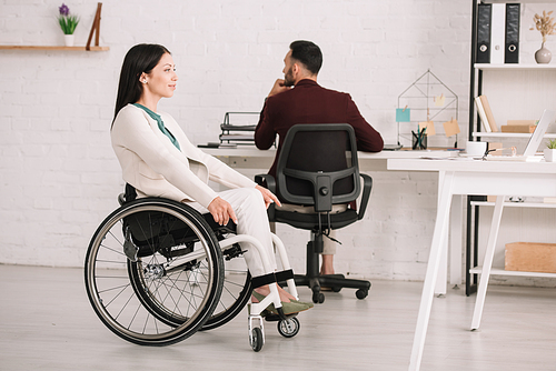 smiling disabled businesswoman looking away while sitting in wheelchair near colleague
