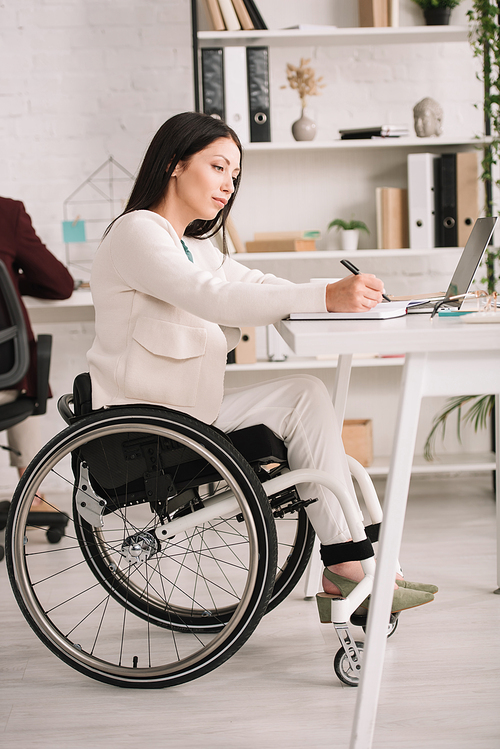 attractive, disabled businesswoman writing in notebook while sitting in wheelchair at workplace