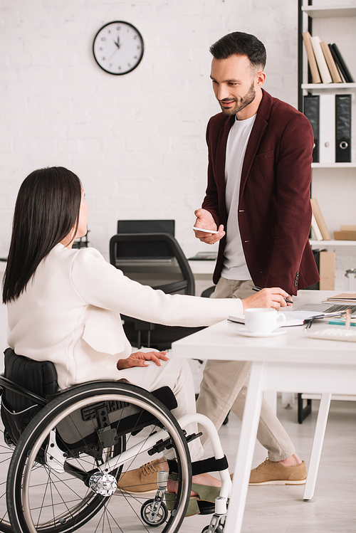 smiling businessman holding smartphone while standing near disabled businesswoman in office