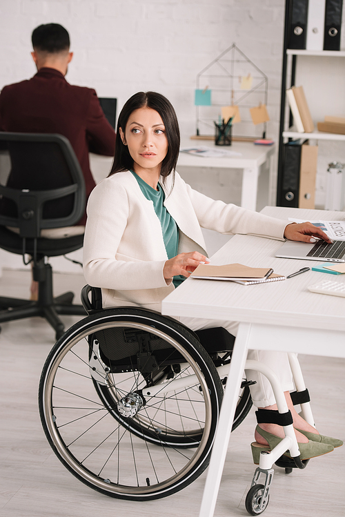 attractive disabled businesswoman looking away while sitting in wheelchair at workplace near colleague on background