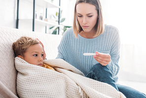 worried woman looking at thermometer while sitting near sick son