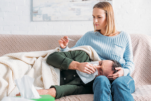 attentive wife looking at thermometer near diseased husband sneezing while lying on her laps