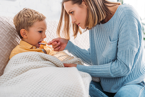 attentive mother giving medicine to diseased son wrapped in blanket
