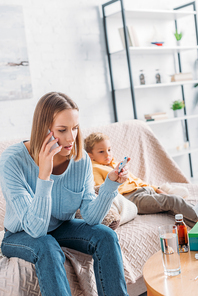 worried woman holding blister of pills and talking on smartphone while sitting near diseased son