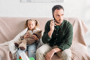 serious man talking on smartphone while sitting near sick daughter