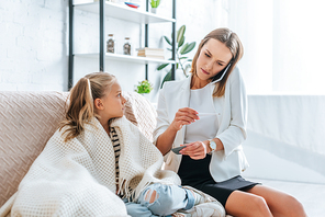 attentive mother talking on smartphone, holding pills and thermometer while sitting near diseased daughter