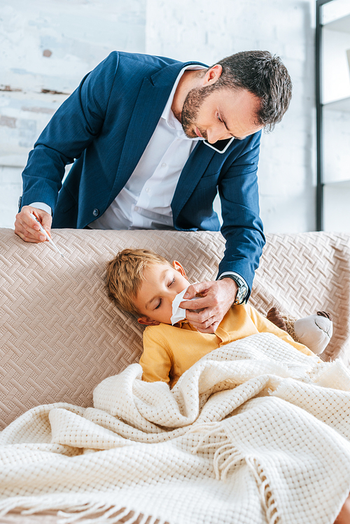 caring father wiping nose of sick son while holding thermometer and talking on smartphone