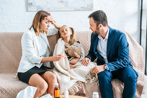 worried mother touching forehead and looking at thermometer while sitting near diseased daughter and husband