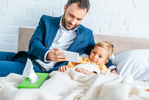 smiling father showing smartphone to sick son lying in bed