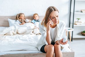 selective focus of upset woman in formal wear talking on smartphone while sitting on bed near sick children and holding container with pills
