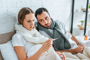 sick woman looking at thermometer near diseased husband
