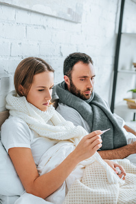 sick woman looking at thermometer near diseased husband