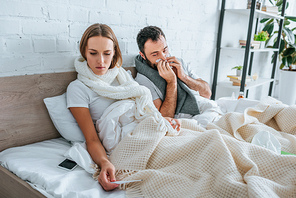 sick woman looking at thermometer near ill husband sneezing in napkin