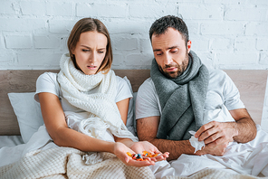 sick woman holding handful of pills while sitting in bed near diseased husband