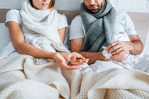 cropped view of sick woman holding handful of pills while sitting in bed near diseased husband