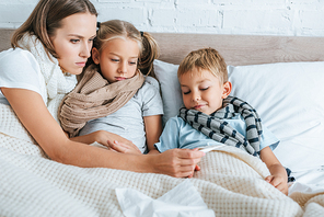 sick mother showing thermometer to diseased children while lying in bed together