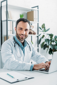 positive doctor looking at camera while using laptop