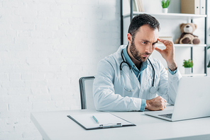 thoughtful doctor looking at laptop while sitting at workplace