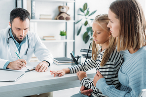 serious doctor writing diagnosis near attentive mother and daughter