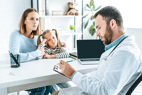 serious pediatrist writing prescription near attentive mother and daughter