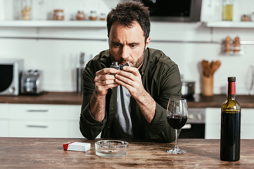 Worried man holding lighter beside wine and cigarettes on kitchen table