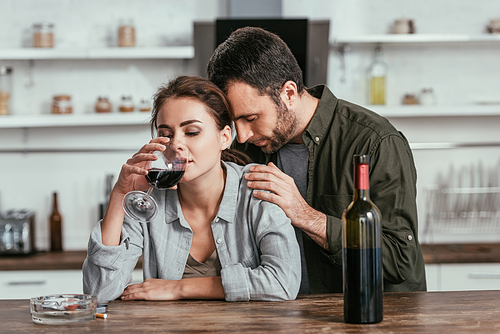 Upset man hugging alcohol addicted wife with wine glass at kitchen