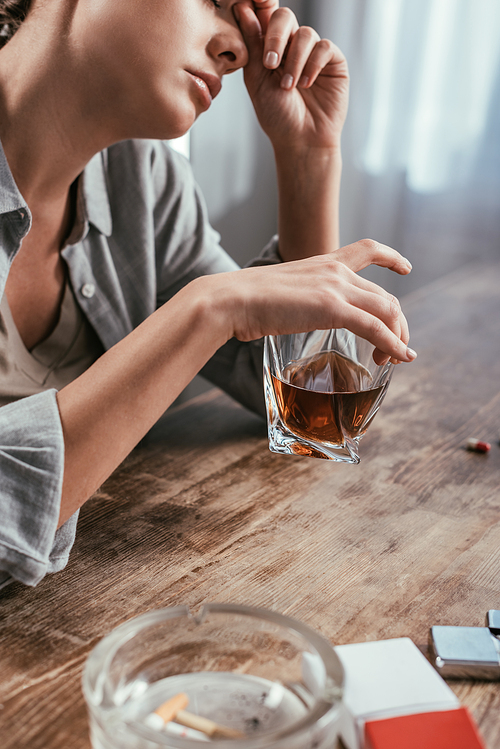 Selective focus of woman with alcohol addiction holding whiskey glass beside cigarettes on table