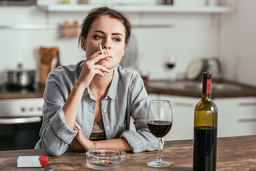 Pensive woman smoking beside wine on kitchen table