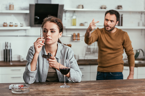 Selective focus of woman with wine smoking while angry husband showing middle finger