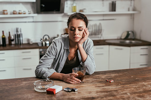 Worried woman holding whiskey glass beside cigarettes and pills on table